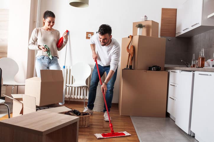 A man mops the floor while a woman holds cleaning supplies. Surrounded by cardboard boxes in the kitchen, the couple prepares to move in and start fresh in their new home.