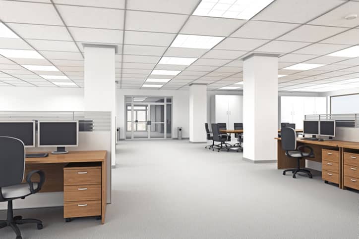 A modern, spacious business office with multiple desks, chairs, and computer monitors arranged in an open layout. The room has bright lighting, white walls, large windows in the background, and highlights the importance of regular carpet cleaning.