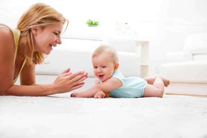 A woman lies on a carpeted floor facing a smiling baby who is on their tummy. The room, recently refreshed after water damage restoration, is bright with a white sofa in the background as the woman claps and smiles.