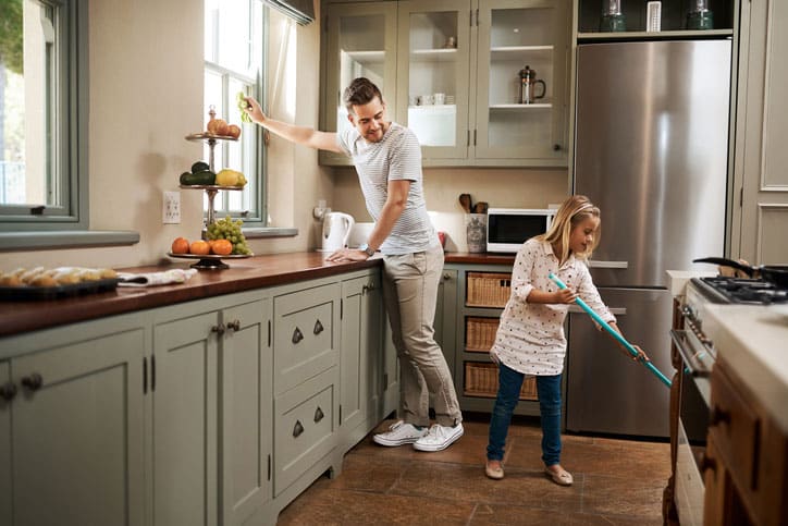 A man opens a kitchen window while a young girl sweeps the floors during Spring cleaning. The kitchen features light green cabinets, stylish furniture, a stainless steel refrigerator, and fruit on the counter. Both appear to be enjoying the chores.