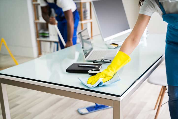 Two people performing office cleaning; one wipes a glass desk with yellow gloves while the other mops in the background. The desk has a laptop, clipboard, and coffee cup, helping to keep the office clean and tidy.