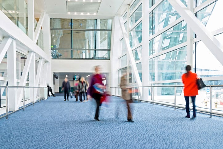 People walk through a bright, modern hallway with large glass windows and white steel beams. The blue carpet, kept fresh with regular commercial carpet cleaning, and natural light create an open, airy atmosphere.