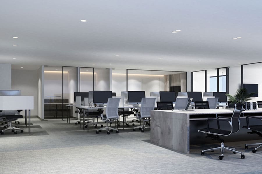 Modern open-plan office with rows of desks, ergonomic chairs, desktop computers, and large windows letting in natural light. The space is clean and spacious, thanks to regular upholstery cleaning that maintains the neutral color palette. No people are present.