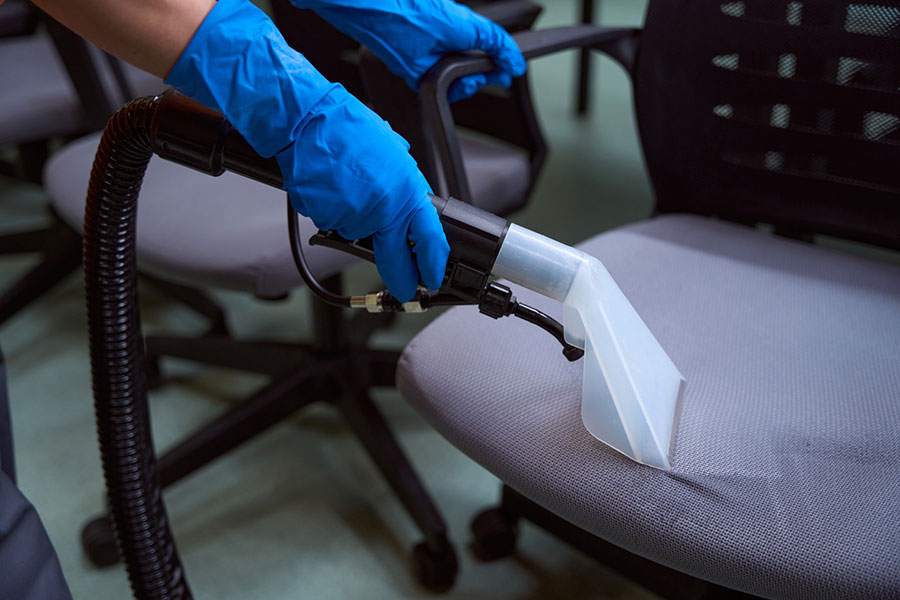 A person wearing blue gloves uses a vacuum extractor to clean the fabric seat of an office chair, surrounded by other empty chairs and various carpet styles in the background.