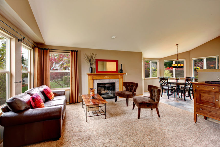 A cozy living room with a brown sofa, two chairs, a wooden coffee table, and a fireplace with a mirror above. Large windows let in natural light. The space is ready for furniture and carpet cleaning to keep it fresh and inviting year-round.