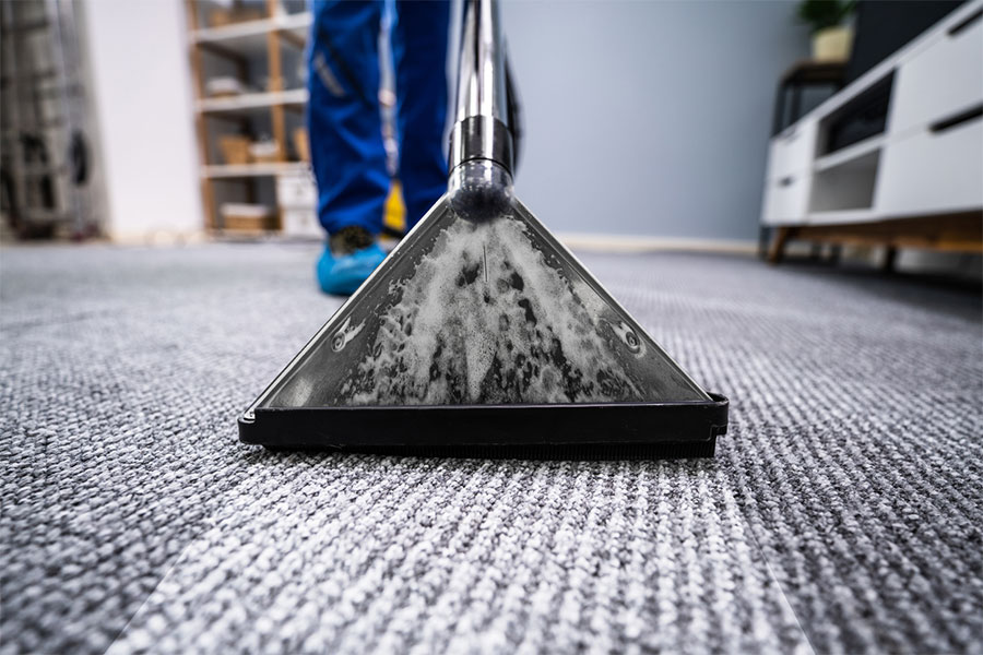 Close-up of a carpet cleaning machine head in action on a gray carpet, highlighting the importance of regular carpet cleaning. A person in blue pants and protective shoe covers works in the background—showcasing how often to clean carpets for best results.