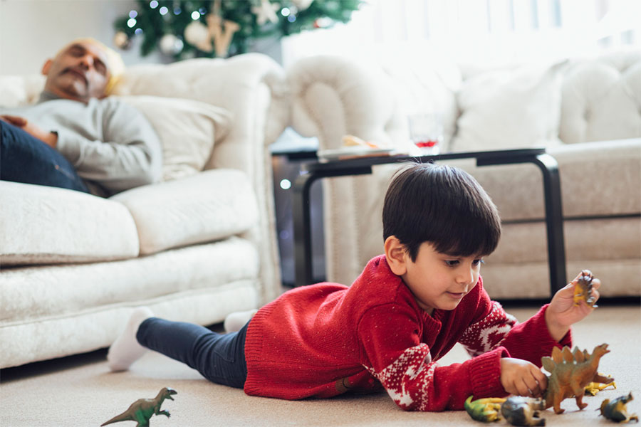 A young boy in a red sweater lies on the carpet playing with toy dinosaurs, while an adult naps on a cream-colored sofa near a decorated Christmas tree—reminding us how important carpet care is during holiday cleaning.